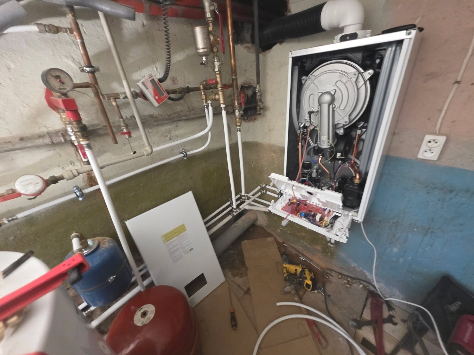 Otwarty piec gazowy z widocznymi elementami wewnętrznymi, rurami i narzędziami w pomieszczeniu technicznym. Widoczne przewody i instalacja.