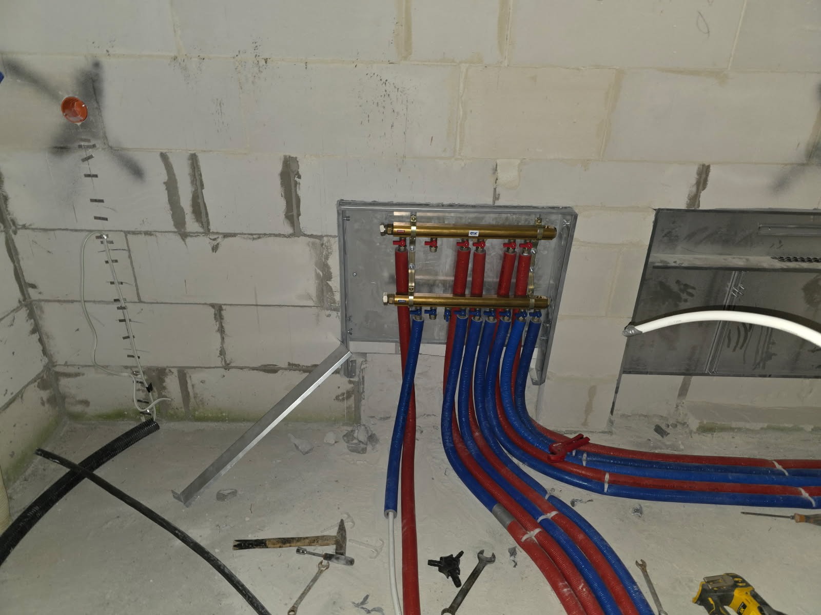 Rozdzielacz ogrzewania podłogowego z rurami PEX w kolorze czerwonym i niebieskim, zamontowany na ścianie z betonu komórkowego. Widoczne narzędzia hydrauliczne.