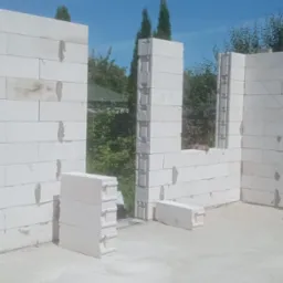 Wznoszenie ścian z białych bloczków betonu komórkowego, widoczne fragmenty ścian z otworami okiennymi i stos bloczków na posadzce, w tle zieleń drzew i błękitne niebo.