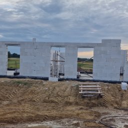 ZA&LE SP.Z.O.O - Wznoszone ściany parterowego domu z białych bloczków na fundamencie, widoczne rusztowanie w otworze okiennym, w tle pola i zachodzące słońce, obok materiały budowlane z logo producenta.