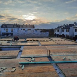 ZA&LE SP.Z.O.O - Budowa domu jednorodzinnego w trakcie realizacji: deskowanie stropu z widocznymi otworami, w tle wznoszone ściany z bloczków SOLBET i zabudowa osiedla w promieniach zachodzącego słońca.