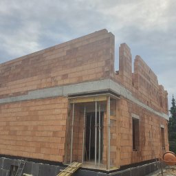 ZA&LE SP.Z.O.O - Wznoszenie ścian zewnętrznych domu z czerwonej cegły ceramicznej, widoczne szalunki okienne i izolacja fundamentów z czarnych płyt, na tle pochmurnego nieba i drzew.