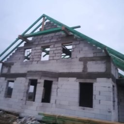 ZA&LE SP.Z.O.O - Wznoszony dwukondygnacyjny budynek z białych bloczków, widoczne otwory okienne i przygotowywana konstrukcja dachu z zielonych belek. Na pierwszym planie ułożone deski.