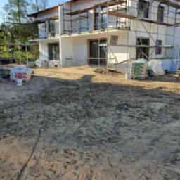 Dwukondygnacyjny dom w trakcie budowy z białych bloczków, otoczony rusztowaniami i materiałami budowlanymi na piaszczystym terenie.