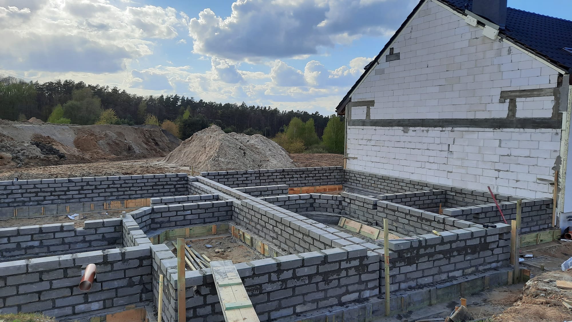 Wykopany i wymurowany fundament pod budynek jednorodzinny, widoczne szalunki i wyprowadzone przyłącze kanalizacyjne, w tle hałda ziemi i las.