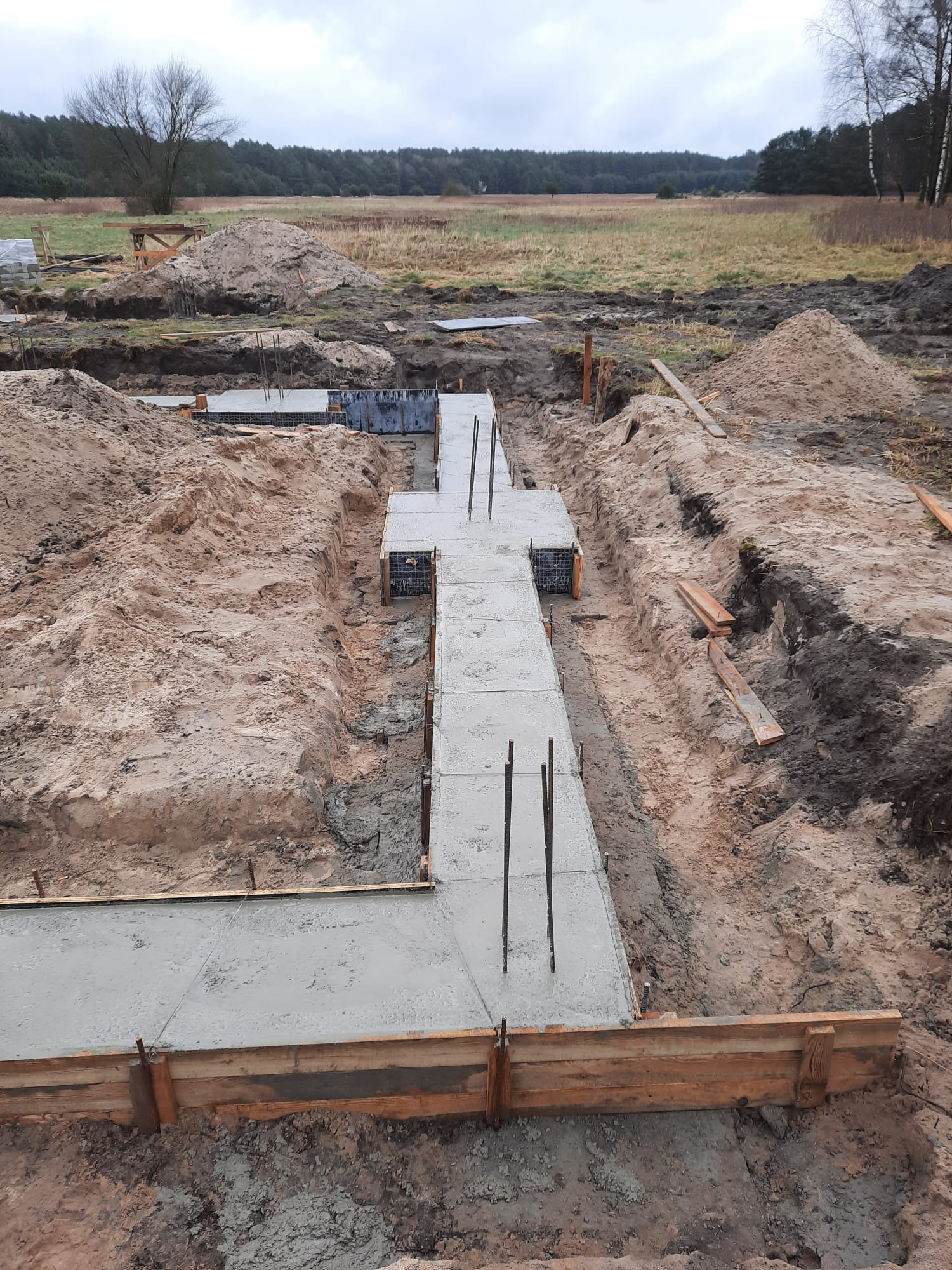 Wylany betonowy fundament z wystającymi prętami zbrojeniowymi w drewnianym szalunku na tle piaszczystego terenu budowy i zalesionego horyzontu pod pochmurnym niebem.