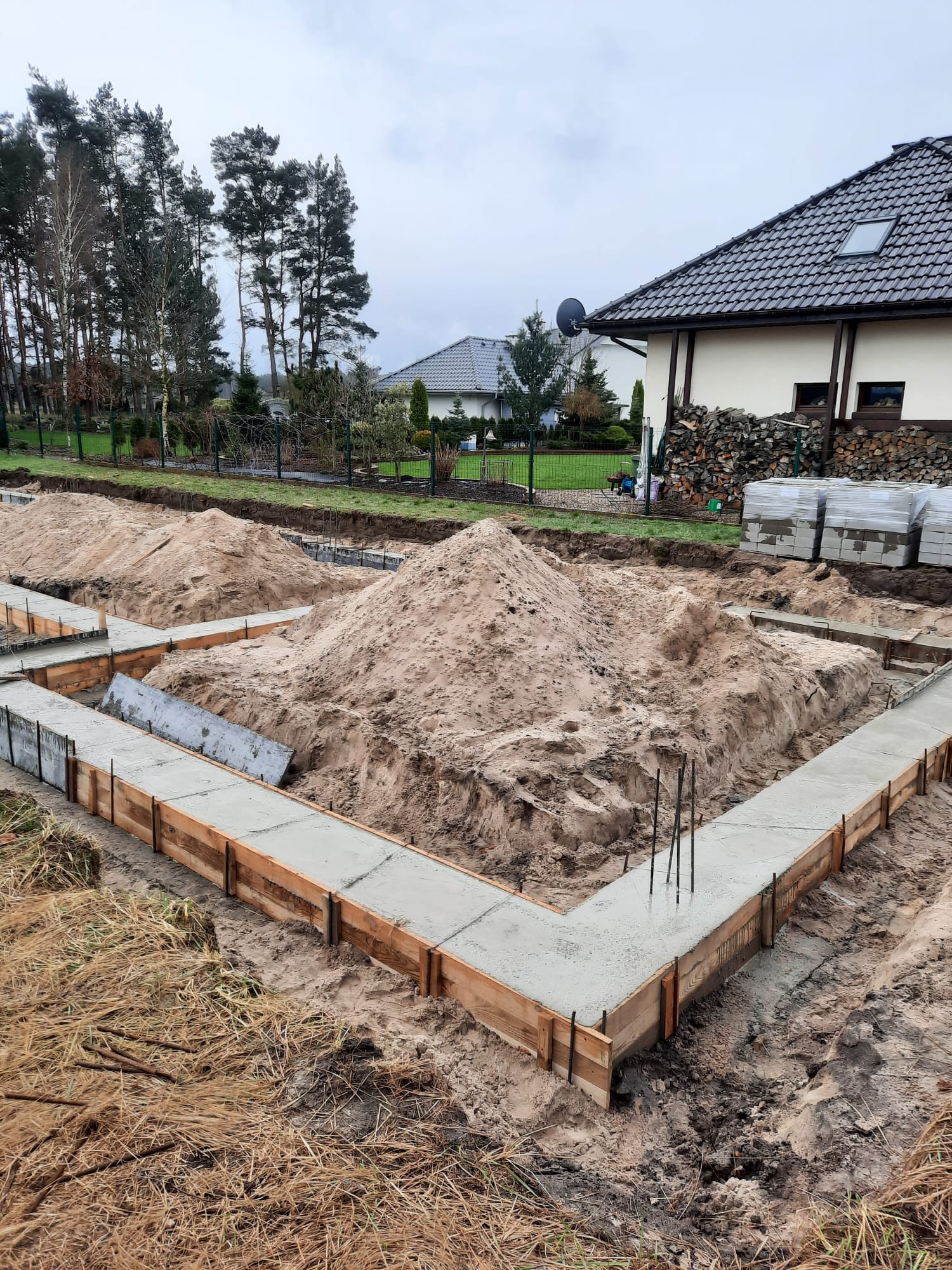 Wylany beton w szalunku fundamentów domu jednorodzinnego, widoczny zbrojenie, w tle sterta piasku, dom z ciemną dachówką i stosem drewna.