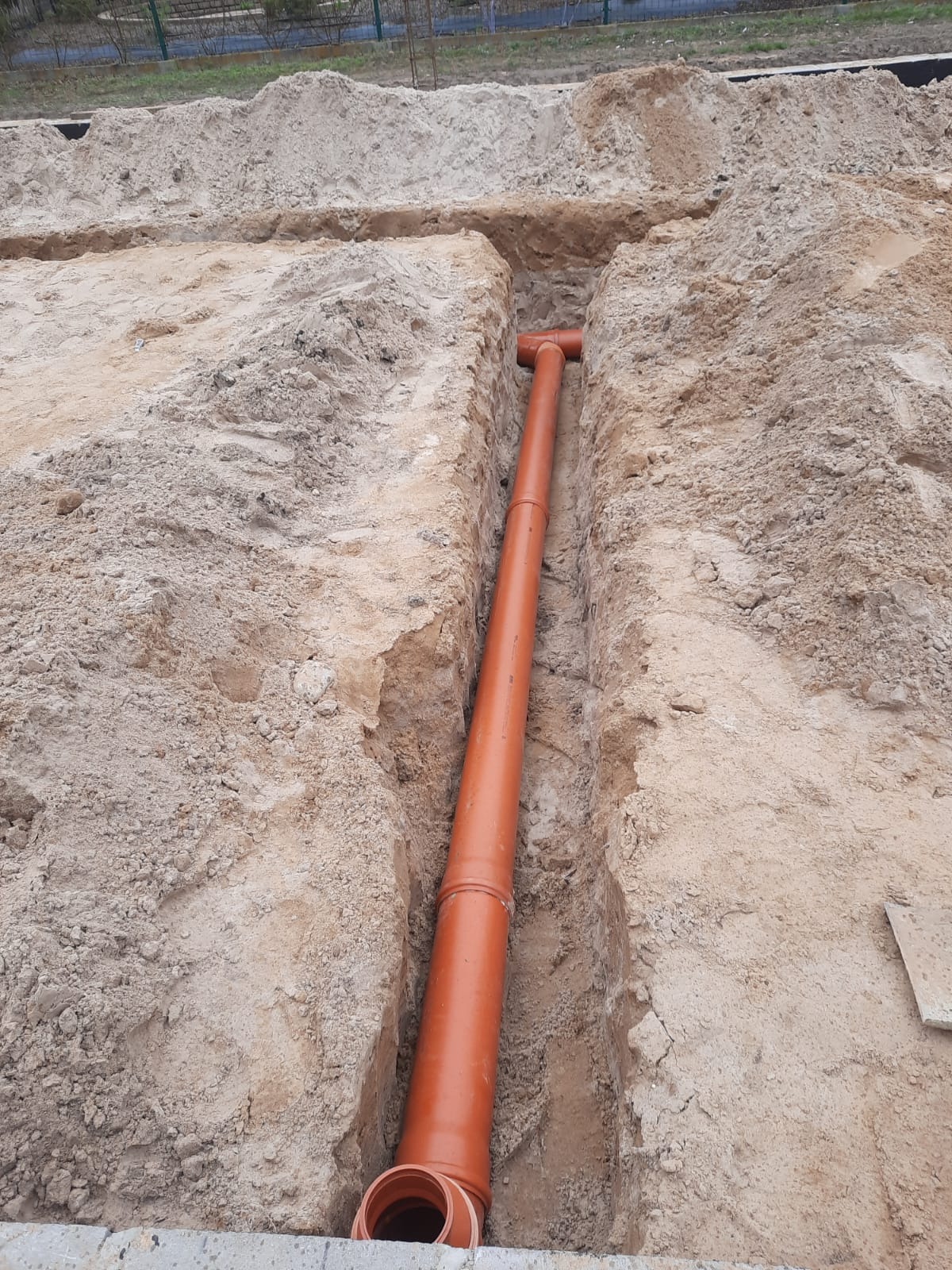 Pomarańczowa rura kanalizacyjna ułożona w wykopie w piaszczystym gruncie, widok z góry ukazuje połączenie trójnikowe i fragment betonowej konstrukcji.