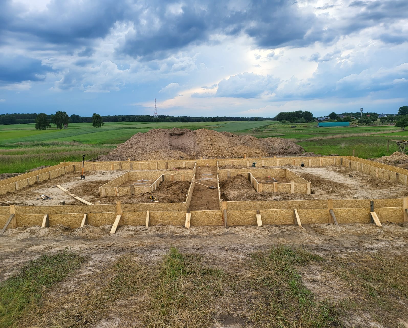 Drewniane szalunki przygotowane pod wylanie fundamentów domu jednorodzinnego na tle pola i pochmurnego nieba.