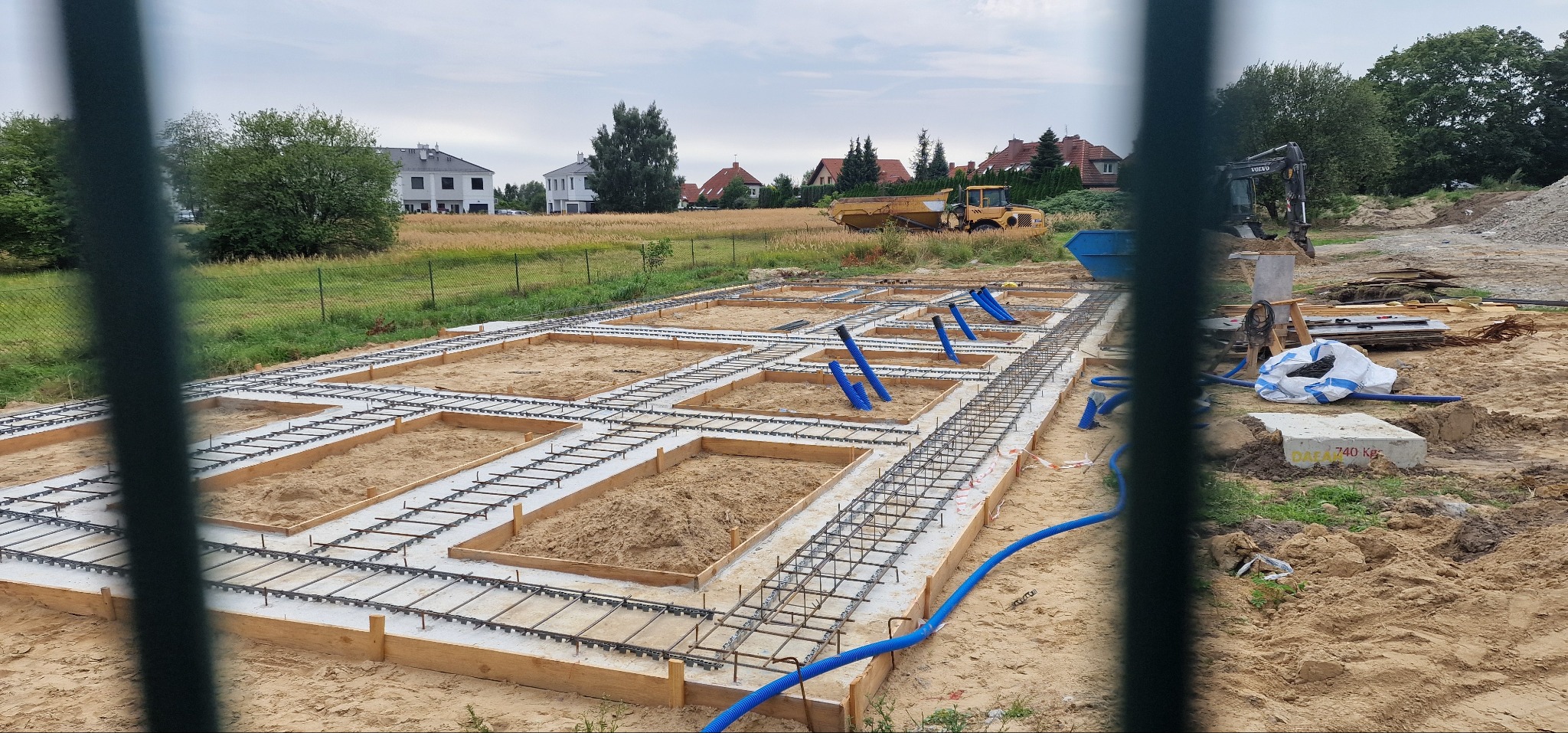 Przygotowanie zbrojenia i szalunku pod płytę fundamentową z widocznymi niebieskimi rurami instalacyjnymi, na tle krajobrazu z domami i maszynami budowlanymi.