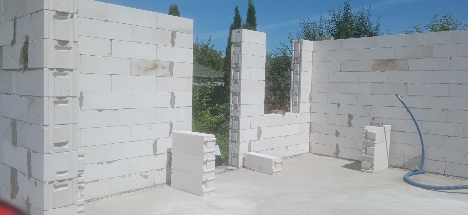 Wznoszenie ścian z białych bloczków betonu komórkowego, widoczne fragmenty ścian z otworami okiennymi i stos bloczków na posadzce, w tle zieleń drzew i błękitne niebo.