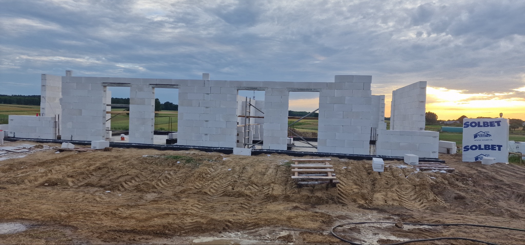 Wznoszone ściany parterowego domu z białych bloczków na fundamencie, widoczne rusztowanie w otworze okiennym, w tle pola i zachodzące słońce, obok materiały budowlane z logo producenta.
