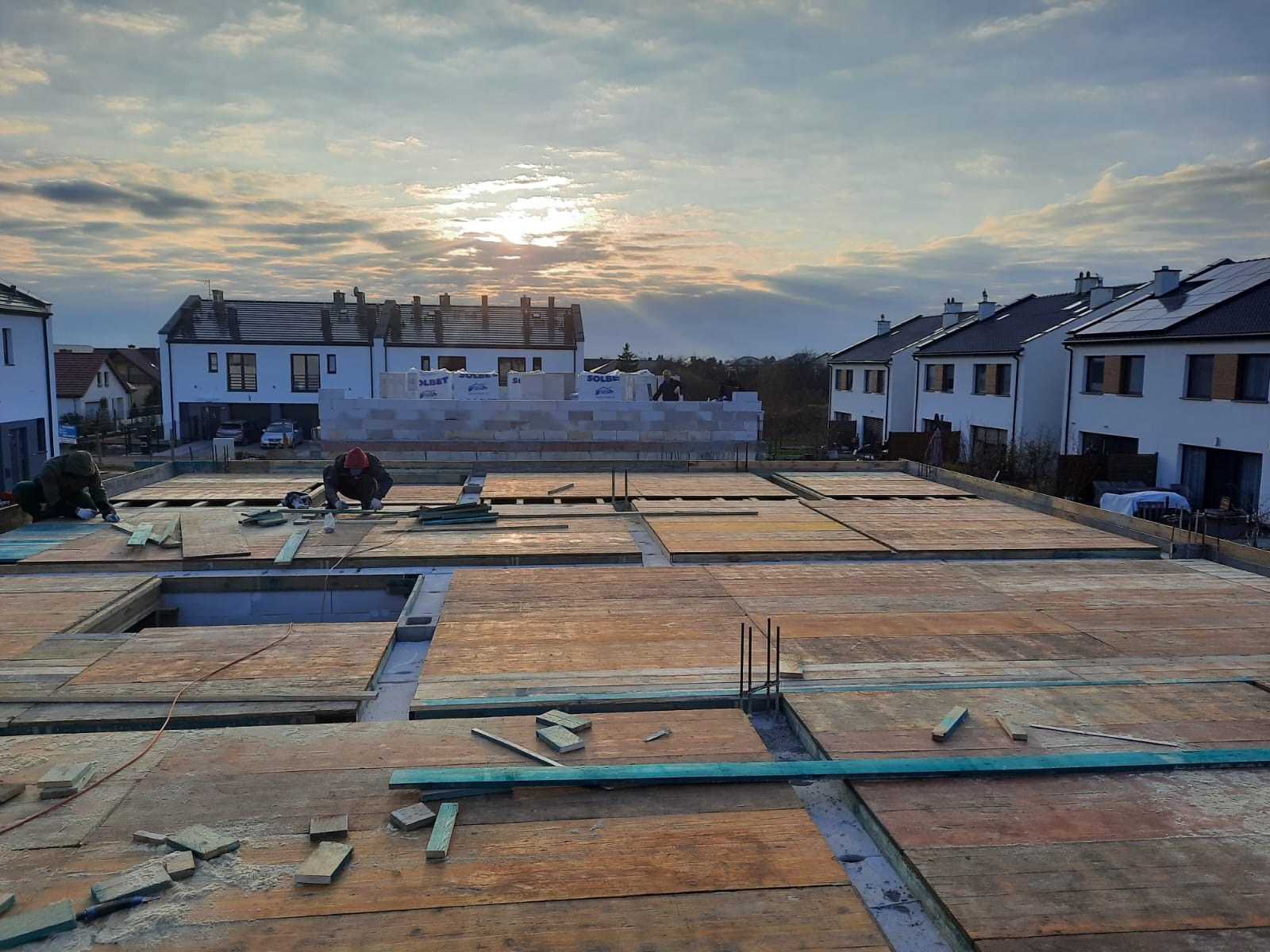 Budowa domu jednorodzinnego w trakcie realizacji: deskowanie stropu z widocznymi otworami, w tle wznoszone ściany z bloczków SOLBET i zabudowa osiedla w promieniach zachodzącego słońca.