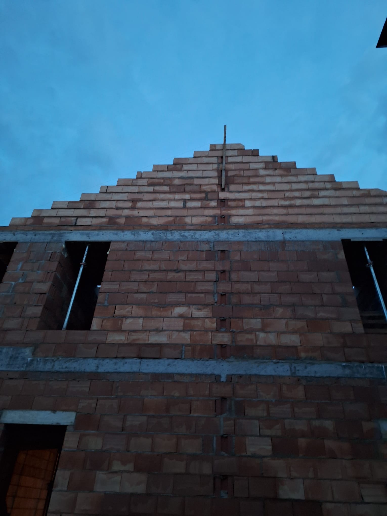 Wznoszenie dwukondygnacyjnego domu z czerwonej cegły ceramicznej w surowym stanie, widoczne otwory okienne ze stalowymi podporami i szczyt dachu na tle pochmurnego nieba.