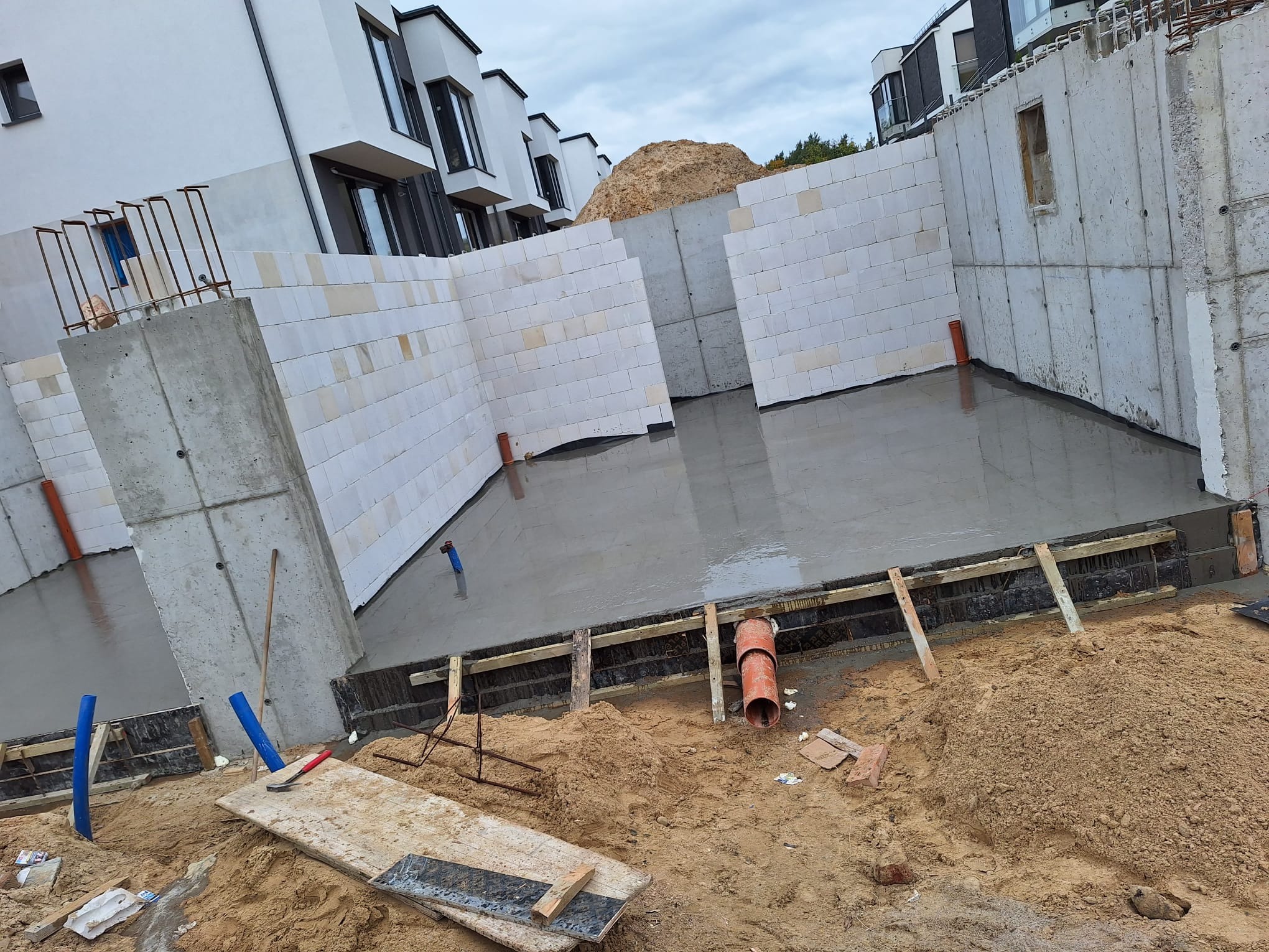 Wylany fundament pod budynek z widocznymi ścianami fundamentowymi z bloczków betonowych i betonu, szalunkami oraz wystającymi rurami instalacyjnymi, na pierwszym planie piasek i narzędzia.