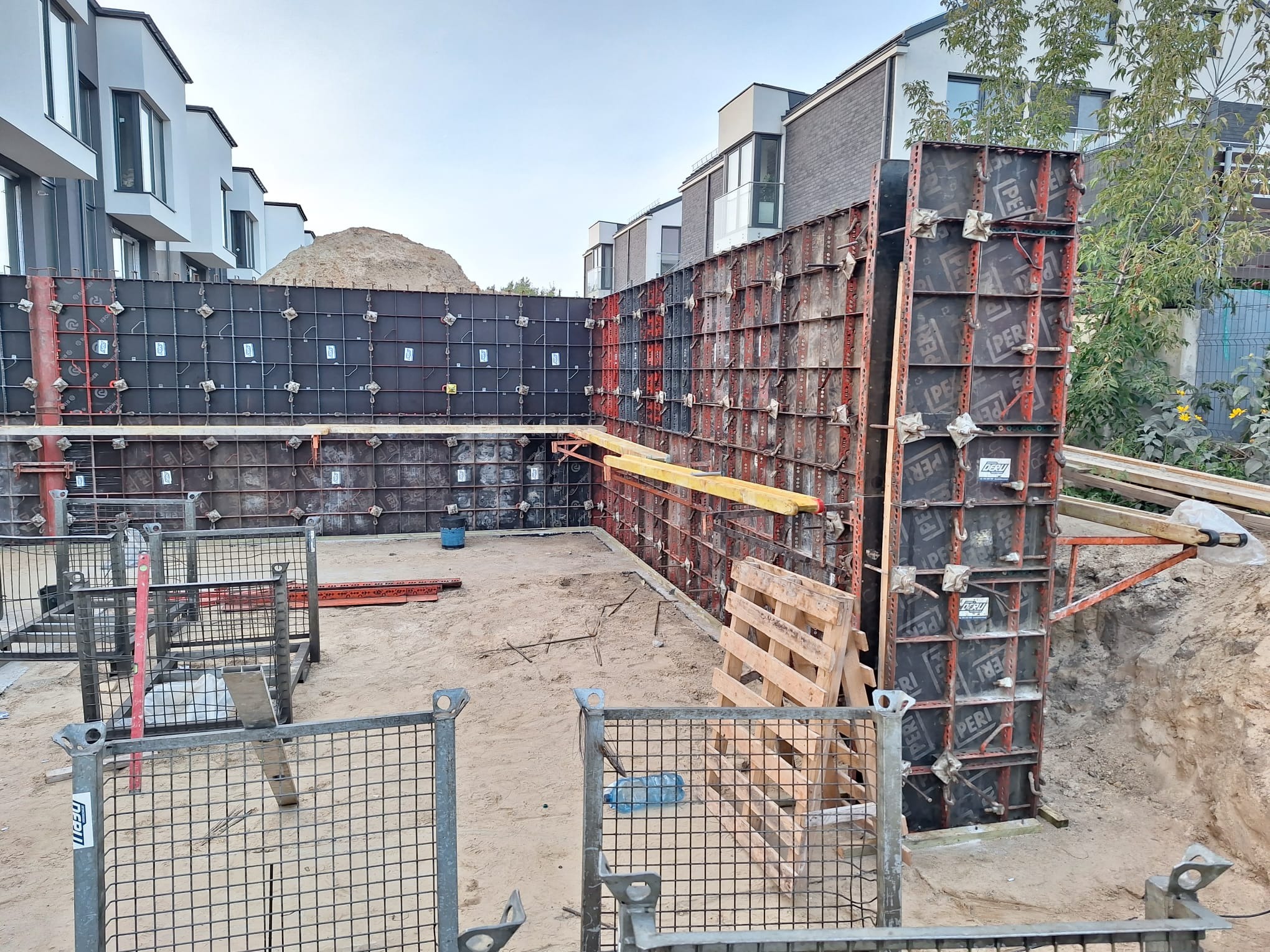 Szare szalunki tworzą kąt prosty na piaszczystym placu budowy, w tle widać nowoczesne budynki mieszkalne i stertę piasku.