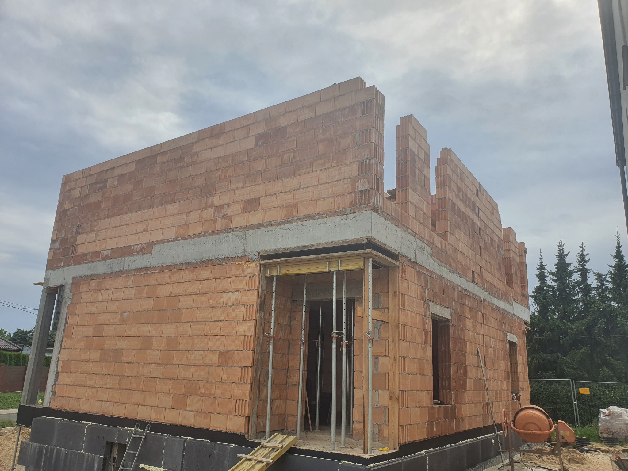 Wznoszenie ścian zewnętrznych domu z czerwonej cegły ceramicznej, widoczne szalunki okienne i izolacja fundamentów z czarnych płyt, na tle pochmurnego nieba i drzew.