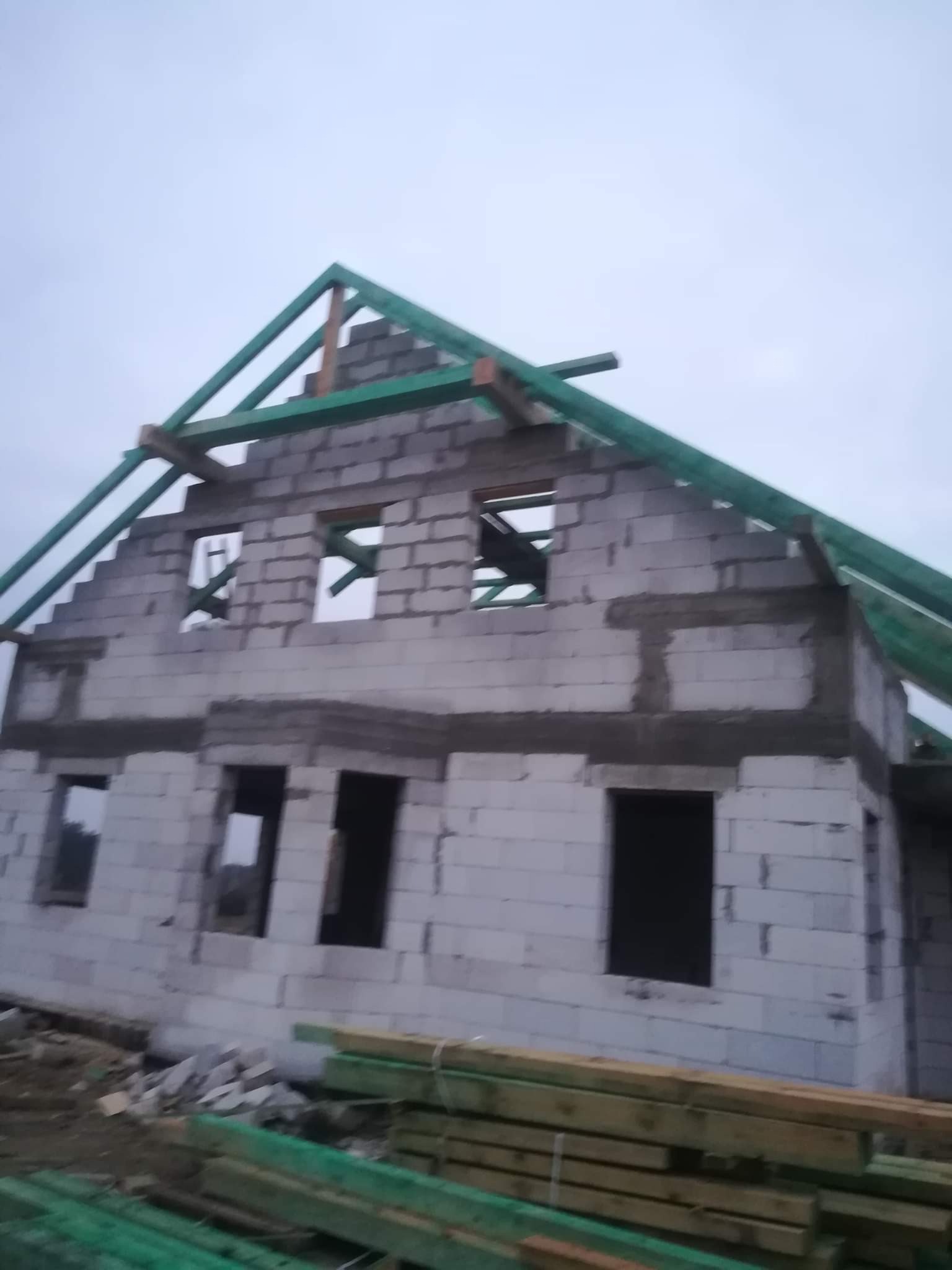 Wznoszony dwukondygnacyjny budynek z białych bloczków, widoczne otwory okienne i przygotowywana konstrukcja dachu z zielonych belek. Na pierwszym planie ułożone deski.