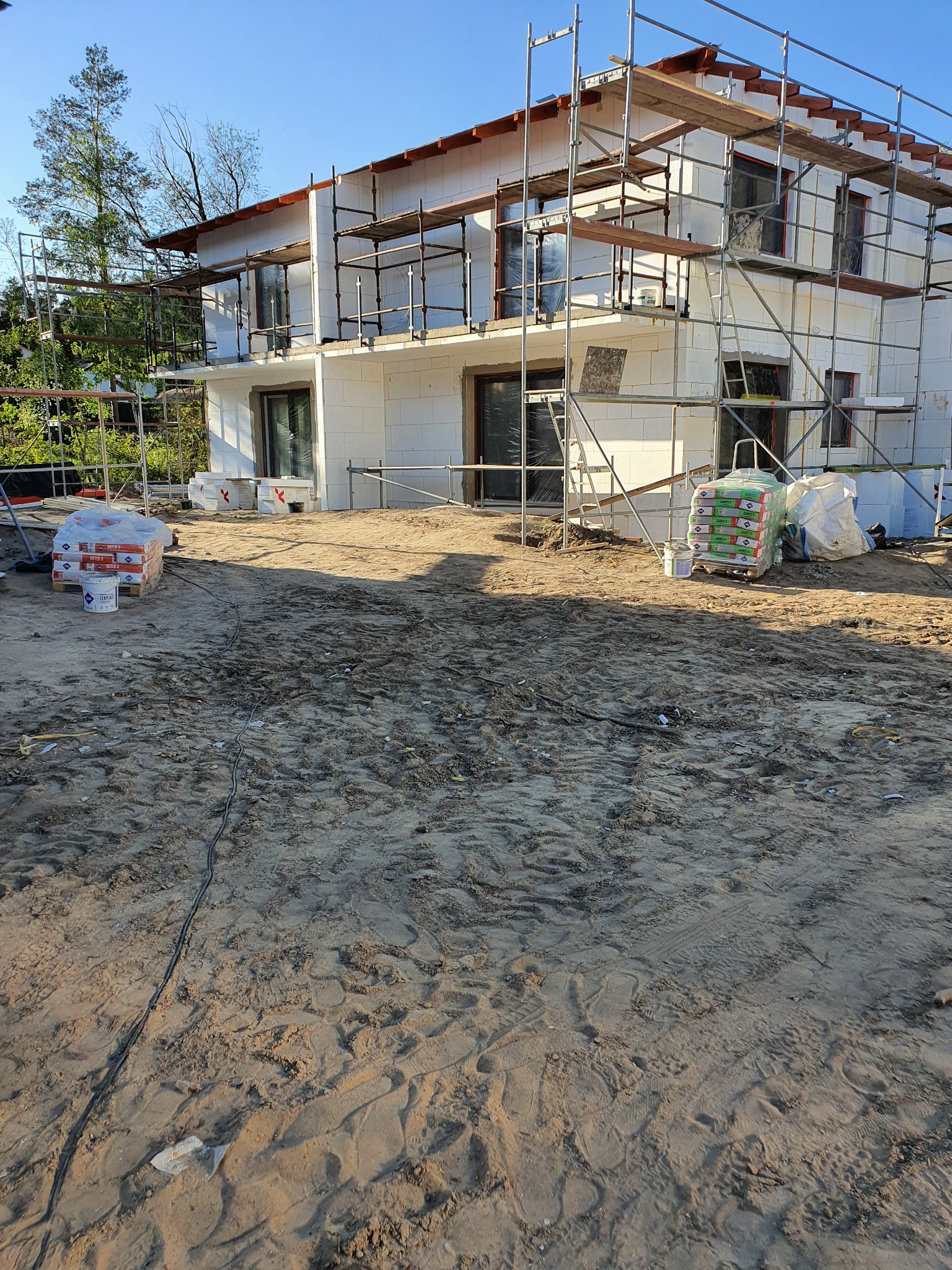 Dwukondygnacyjny dom w trakcie budowy z białych bloczków, otoczony rusztowaniami i materiałami budowlanymi na piaszczystym terenie.