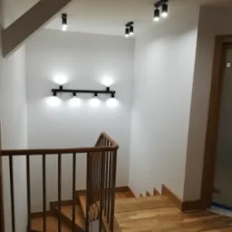 Klatka schodowa z drewnianymi schodami i balustradą, oświetlona punktowymi lampami sufitowymi i listwą LED na ścianie, wykończona w jasnych barwach.