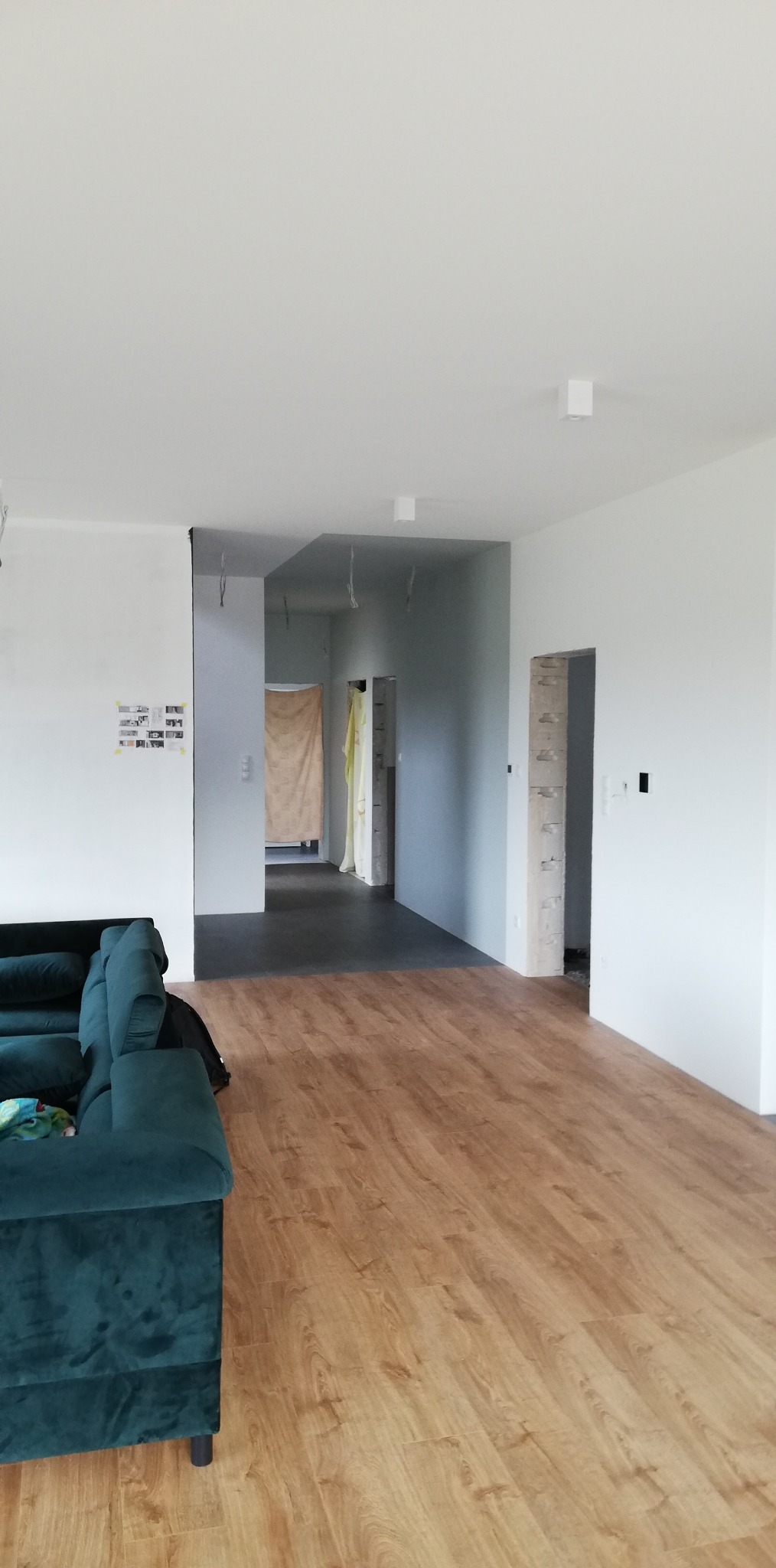 Wnętrze w trakcie remontu: świeżo położona podłoga laminowana w odcieniach drewna, surowe ściany z widocznymi instalacjami elektrycznymi, częściowo zakryte przejście folią malarską...