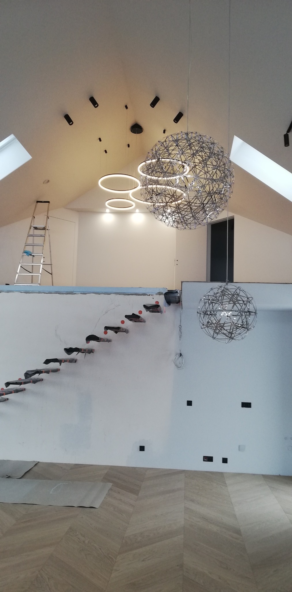 Wnętrze w trakcie prac wykończeniowych: schody ze stopniami zabezpieczonymi folią, podłoga w jodełkę, nowoczesne lampy wiszące i sufitowe, drabina oparta o ścianę.