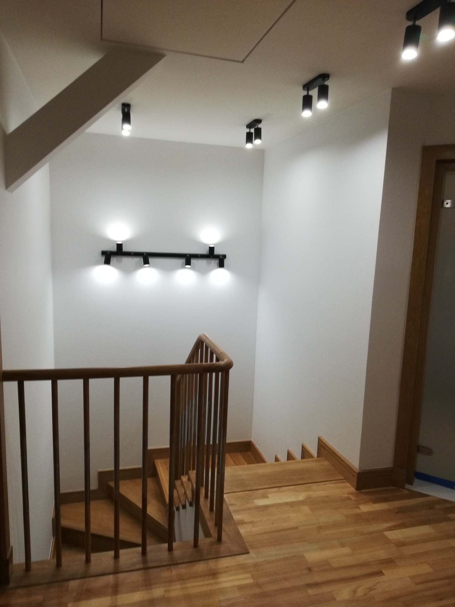Klatka schodowa z drewnianymi schodami i balustradą, oświetlona punktowymi lampami sufitowymi i listwą LED na ścianie, wykończona w jasnych barwach.