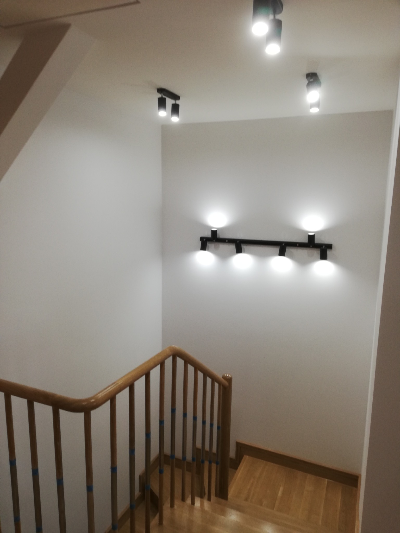 Klatka schodowa z drewnianymi schodami i balustradą, oświetlona nowoczesnymi reflektorami sufitowymi i ściennymi.