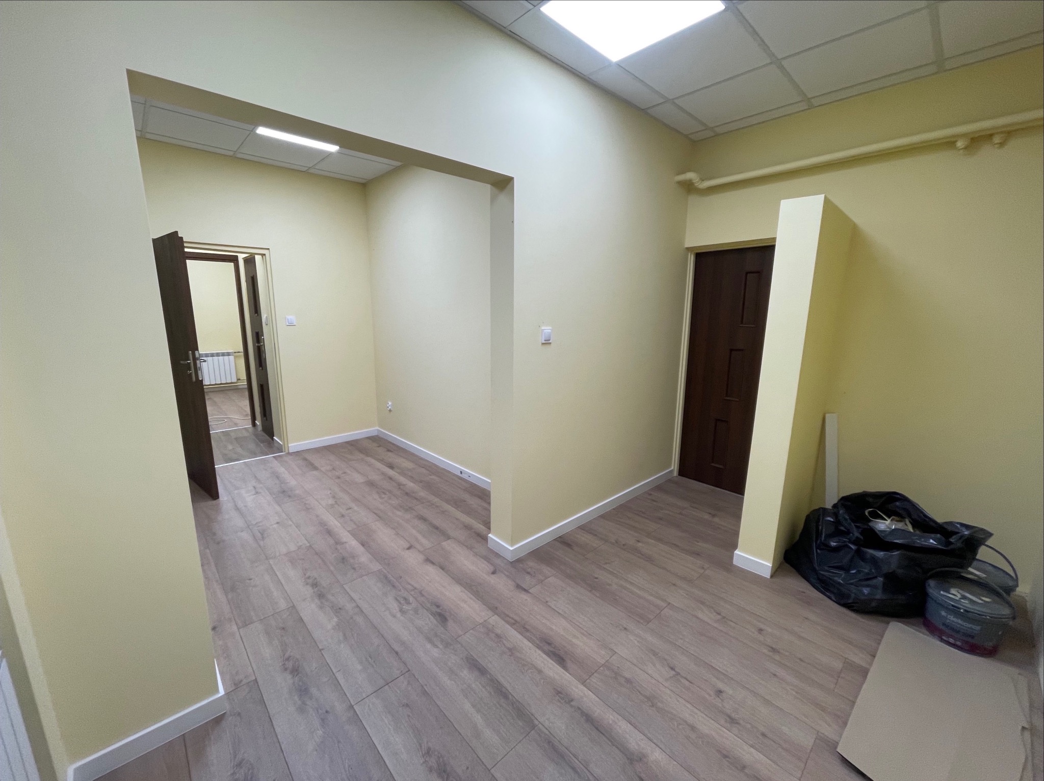 Jasne wnętrze po remoncie: świeżo pomalowane ściany w odcieniach beżu, panele podłogowe imitujące drewno, białe listwy przypodłogowe, widoczne drzwi i fragmenty sufitu.