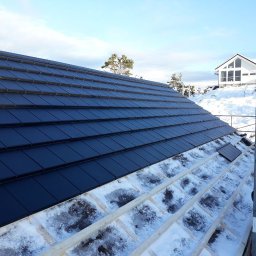 Krystian Wójs DACHYWOJS - Częściowo pokryty dach z czarnych dachówek w zimowej scenerii. Widoczna więźba dachowa pokryta śniegiem i fragment białego domu w tle.