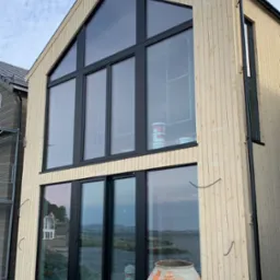 Surowy, dwukondygnacyjny dom modułowy z jasnego drewna, z dużymi oknami i widokiem na wodę. Na pierwszym planie betoniarka i elementy rusztowania.