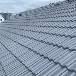 Ukończony dach z szarej dach&oacute;wki ceramicznej, widok z g&oacute;ry. W tle dom z białymi ścianami i ciemnym dachem, częściowo widoczne rusztowanie.