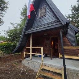 Krystian Wójs DACHYWOJS - Drewniany domek letniskowy w stylu A-frame z ciemnym, profilowanym pokryciem dachowym i polską flagą na szczycie. Widoczne są niedokończone drewniane schody i balustrada tarasu.