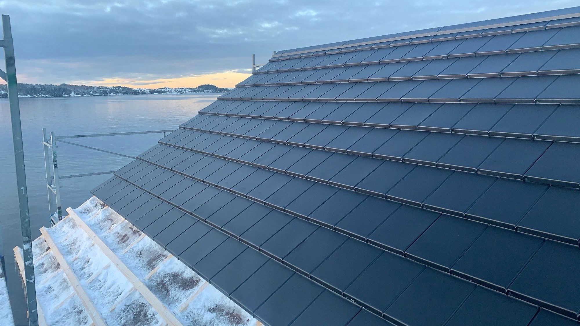 Nowy dach z ciemnych dachówek ceramicznych, częściowo pokryty śniegiem. W tle widok na jezioro i zachodzące słońce. Elementy rusztowania po lewej stronie kadru.