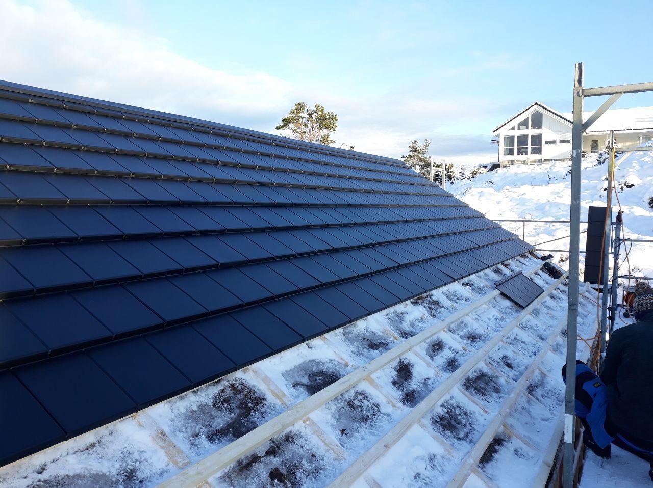 Częściowo pokryty dach z czarnych dachówek w zimowej scenerii. Widoczna więźba dachowa pokryta śniegiem i fragment białego domu w tle.