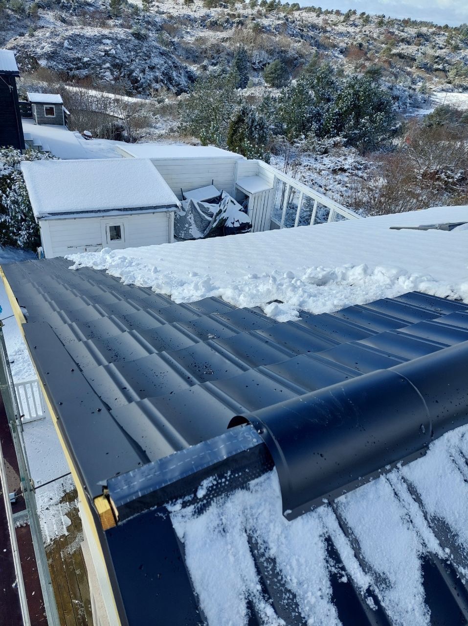 Montaż czarnej blachy na dachu w zimowym krajobrazie. Widoczne zabezpieczenia i fragment ośnieżonego otoczenia. Dach pokryty częściowo śniegiem.