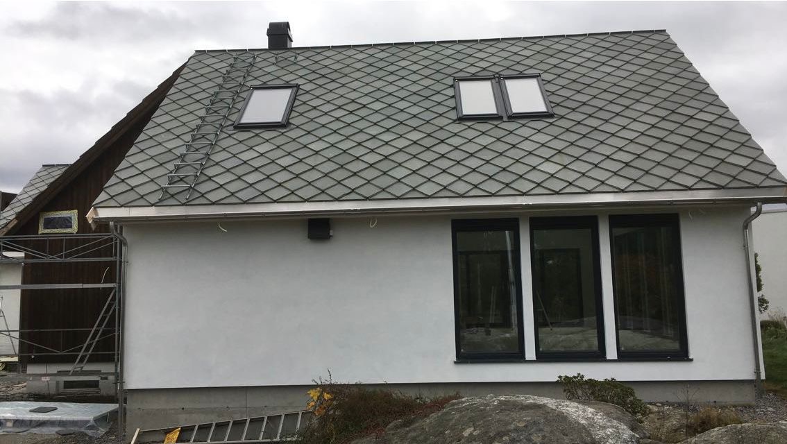 Dom z dachem krytym szarą dachówką rombową, dwa okna dachowe i drabina oparta o dach. Widoczne trzy okna w ścianie bocznej budynku i fragment rusztowania.