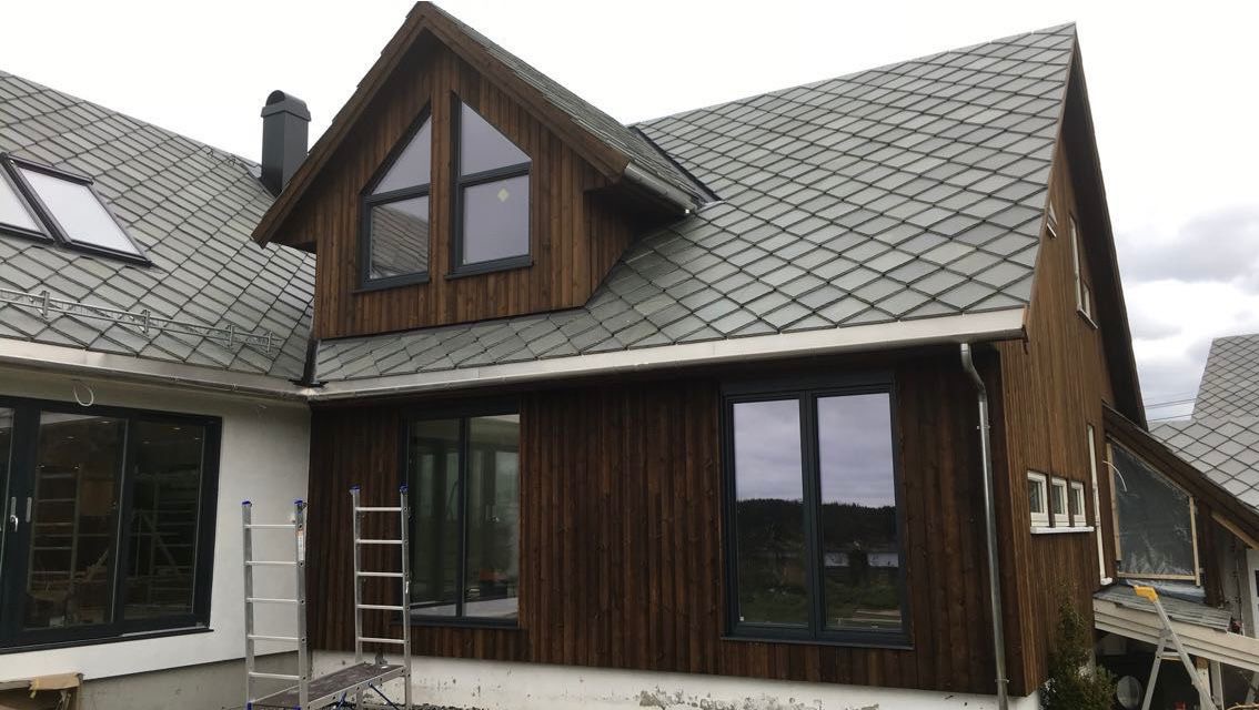 Nowoczesny dom z drewnianą elewacją i szarym dachem z dachówki karpiówki. Widoczne okna z ciemnymi ramami i drabina oparta o ścianę. Detal: charakterystyczne okna na poddaszu.