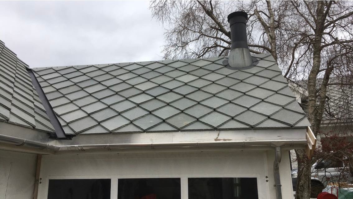 Szary dach z dachówki rombowej z kominem i rynną, widok z dołu na tle drzewa bez liści i pochmurnego nieba. Charakterystyczny wzór ułożenia.