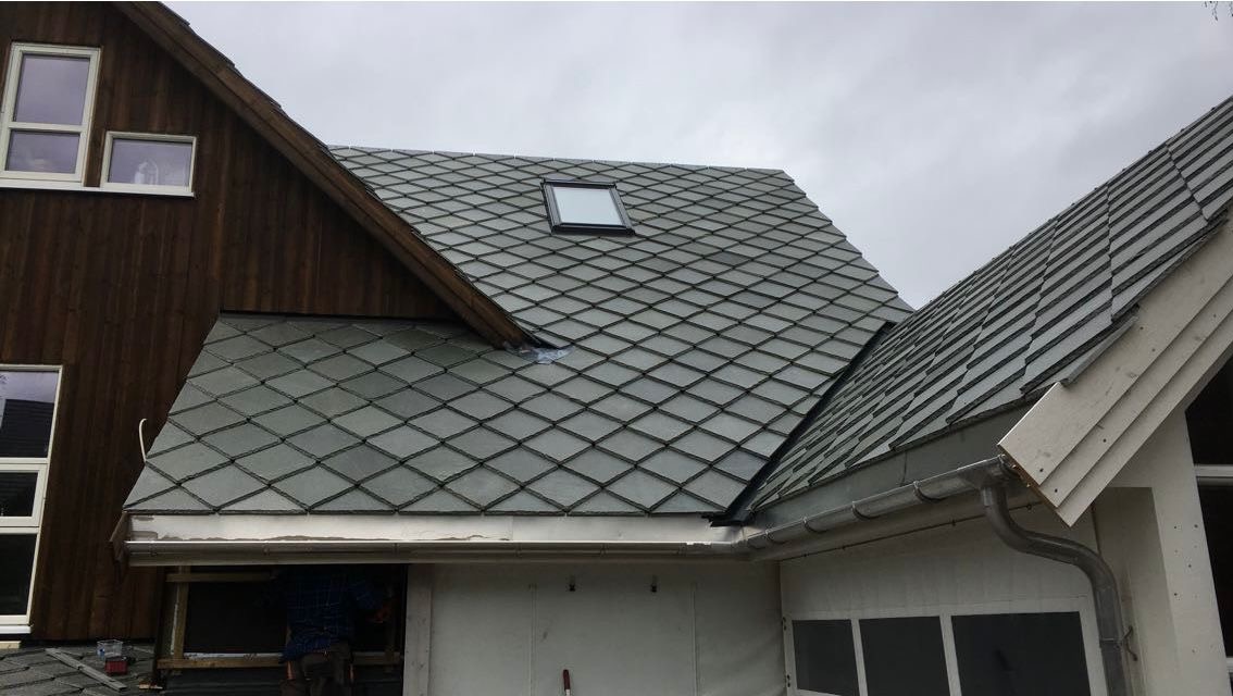 Szary dach z dachówką rombową i oknem dachowym. Fragment drewnianej ściany budynku z oknami. Montaż orynnowania.