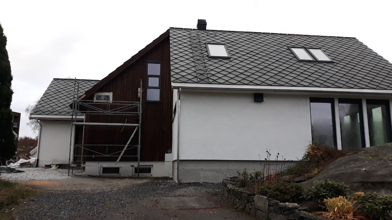 Dom jednorodzinny z ciemną drewnianą elewacją i dachem z dachówki karpiówki, na którym widoczna jest drabinka dachowa. Część domu jest otynkowana na biało.