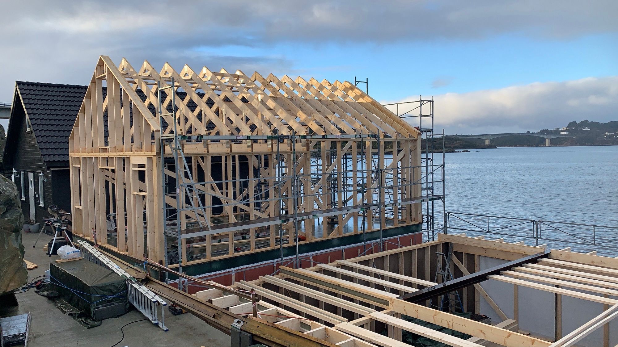 Wznoszenie drewnianej konstrukcji domu modułowego z widokiem na wodę i most w tle. Widoczna więźba dachowa i fragmenty rusztowania.