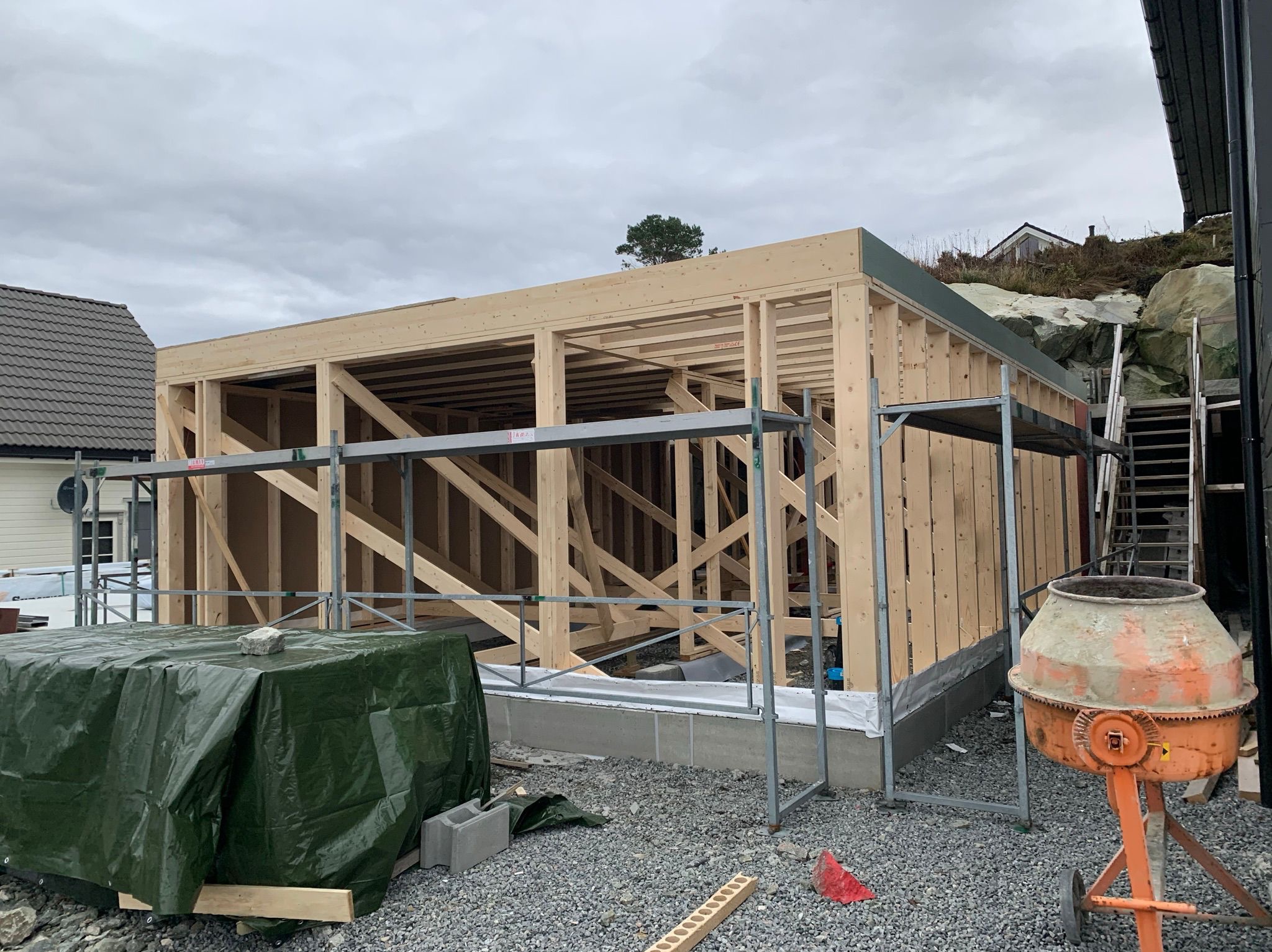 Surowa konstrukcja domu modułowego z drewna, z widoczną ramą i elementami szkieletowymi. W tle zielony krajobraz i pochmurne niebo.