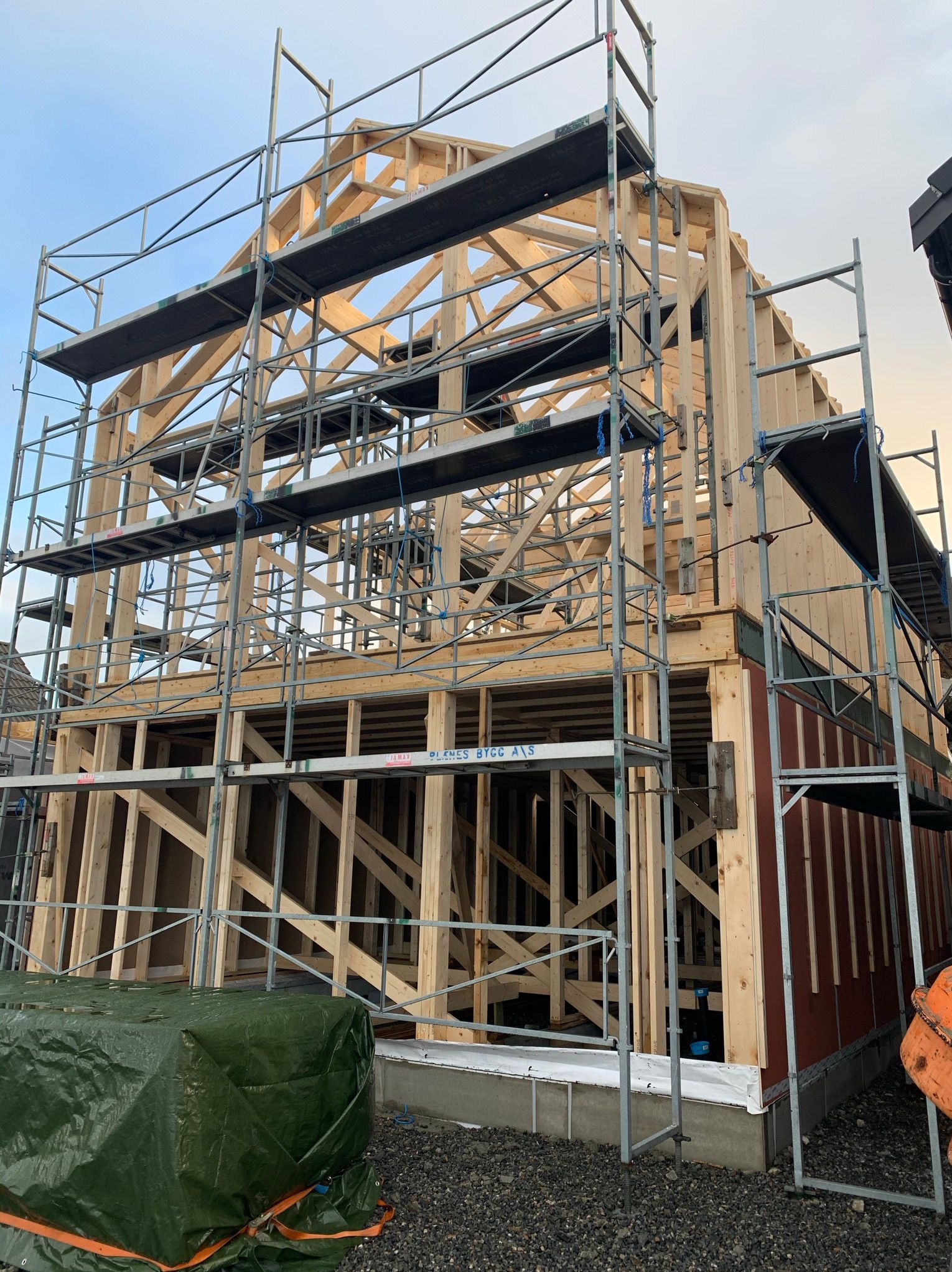 Surowa drewniana konstrukcja domu modułowego w trakcie budowy, otoczona metalowymi rusztowaniami. Widoczne elementy ścian i dachu, przygotowane do dalszych prac.