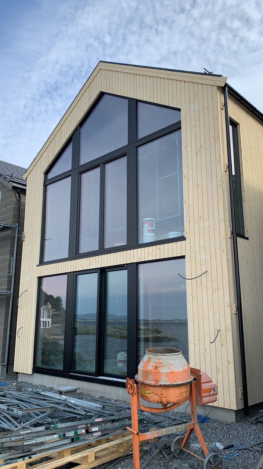 Surowy, dwukondygnacyjny dom modułowy z jasnego drewna, z dużymi oknami i widokiem na wodę. Na pierwszym planie betoniarka i elementy rusztowania.