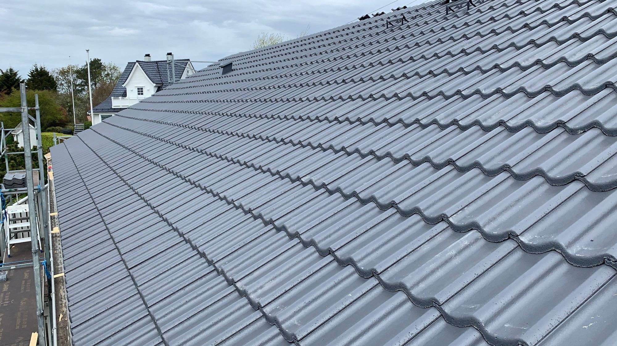 Ukończony dach z szarej dachówki ceramicznej, widok z góry. W tle dom z białymi ścianami i ciemnym dachem, częściowo widoczne rusztowanie.