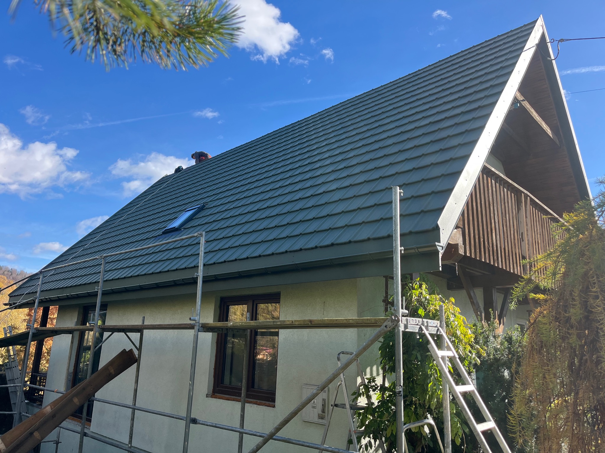 Dom z nowym, grafitowym pokryciem dachowym z dachówki, widoczny z rusztowania. Ujęcie z dołu, błękitne niebo w tle, fragment iglastego drzewa w rogu.