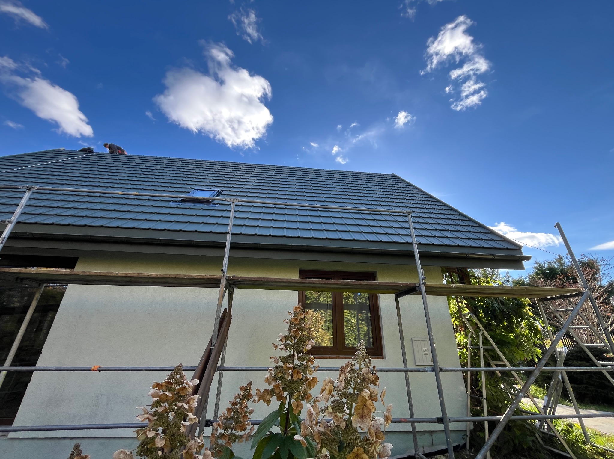 Dom jednorodzinny z nowym, grafitowym pokryciem. Na dachu widoczny dekarz podczas pracy. Rusztowanie przy ścianie budynku. Błękitne niebo z chmurami w tle.