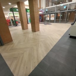 ABRAM-BUD WORTHY - Jasna, drewnopodobna podłoga ułożona w jodełkę w przestrzeni komercyjnej, kontrastująca z ciemnymi płytkami. Widoczne filary i wejście do sklepu.