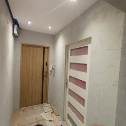 ABRAM-BUD WORTHY - Korytarz w trakcie prac: ściany pokryte szarą strukturą, widoczne dwie różne ościeżnice drzwiowe, zabezpieczona podłoga i wiadro z narzędziem.